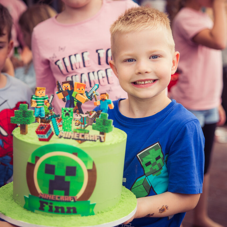 Junge mit Minecraft-Geburtstagstorte, strahlendes Lächeln, fröhliche Kinderparty im Hintergrund
