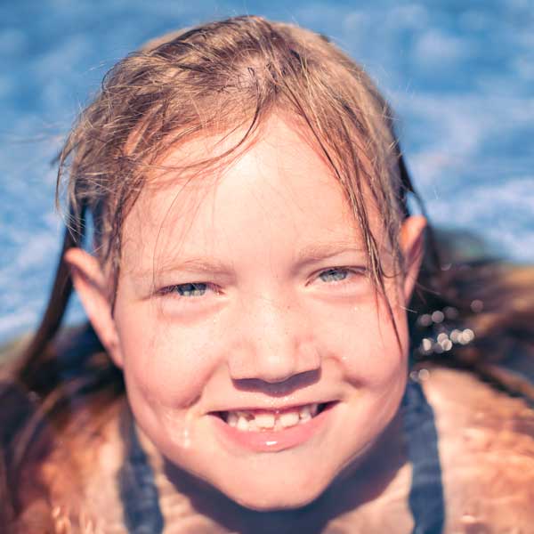Mädchen mit nassen Haaren lächelt fröhlich im Pool in die Kamera, sommerliches Kinderportrait