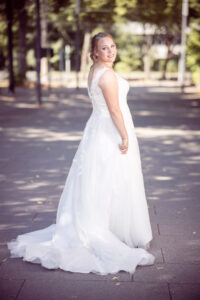 Eine Braut im weißen Spitzenkleid dreht sich auf einem sonnigen Weg über die Schulter zur Kamera und lächelt. Ihr Kleid fällt weich über den Boden, das Licht spielt mit den Schatten der Bäume – ein natürlicher Hochzeitsmoment voller Wärme.