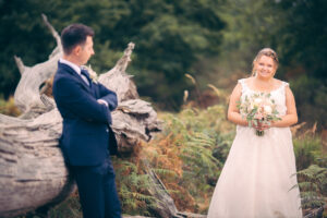 Eine Braut steht mit Blumenstrauß vor einem gefallenen, knorrigen Baumstamm in einem naturbelassenen Areal. Sie schaut liebevoll zu ihrem Bräutigam, der unscharf im Vordergrund am Baum lehnt. Der Moment wirkt ruhig, natürlich und voller Zuneigung.