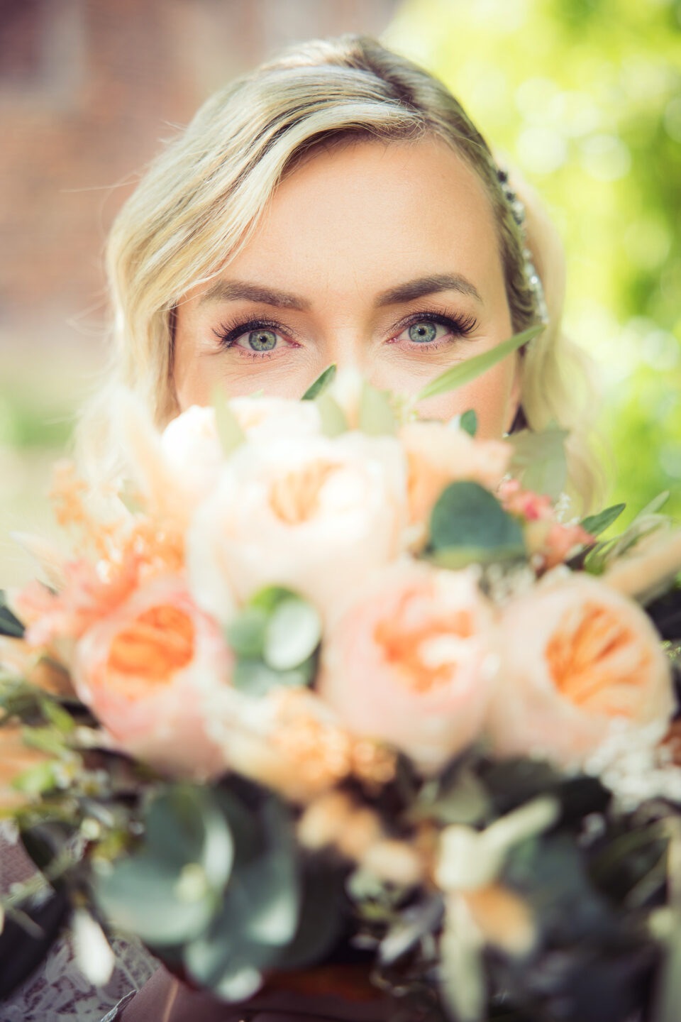 Nahaufnahme einer Braut, die durch ihren pastellfarbenen Blumenstrauß blickt – Fokus auf ausdrucksstarken grünen Augen und sanftem Bokeh im Hintergrund.