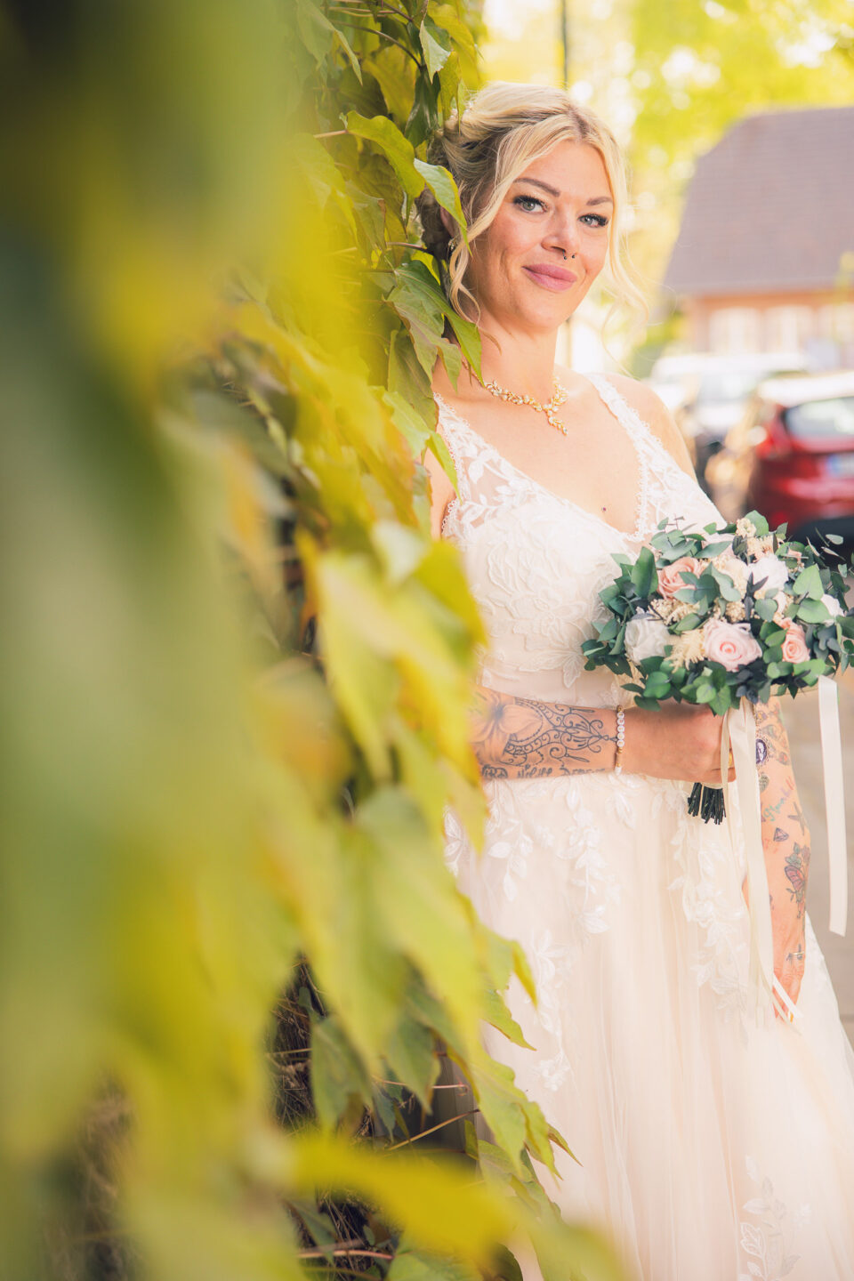 Eine tätowierte Braut lehnt mit einem natürlichen Lächeln an einer mit grünen Blättern bewachsenen Wand. Sie trägt ein elegantes, besticktes Brautkleid und hält einen Blumenstrauß mit Rosen und Eukalyptus.