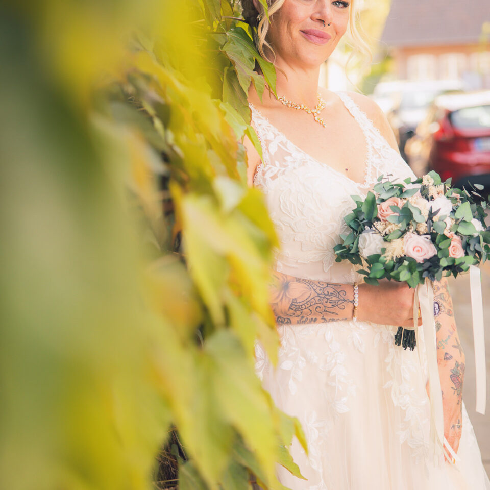 Eine tätowierte Braut lehnt mit einem natürlichen Lächeln an einer mit grünen Blättern bewachsenen Wand. Sie trägt ein elegantes, besticktes Brautkleid und hält einen Blumenstrauß mit Rosen und Eukalyptus.