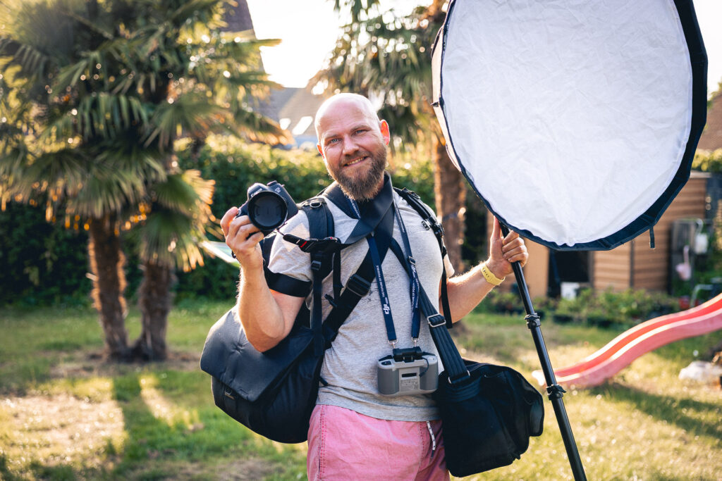 Fotograf Dominik Tetzlaff mit umfangreichem Fotoequipment, Softbox und Kamera, bereit für ein Shooting im Garten – humorvoll inszeniert.