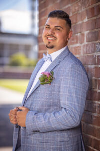 Ein Bräutigam lehnt entspannt an einer Backsteinwand, trägt einen hellblauen Anzug mit Fliege und einem floralen Anstecker. Er lacht in die Kamera – ein natürlicher, authentischer Hochzeitsmoment.