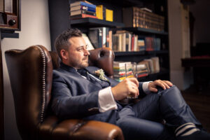 Ein Bräutigam sitzt entspannt im Ledersessel einer Bibliothek, hält ein Glas in der Hand und blickt nachdenklich zur Seite. Die Szene ist stilvoll und ruhig – ein Moment der Vorfreude vor der Trauung.