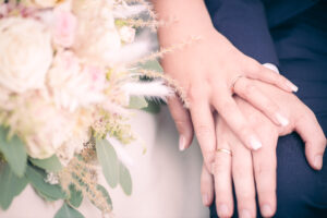 Detailaufnahme von zwei sich berührenden Händen mit Eheringen – im Vordergrund ein pastellfarbener Hochzeitsstrauß mit Rosen, Eukalyptus und Gräsern. Sanftes Licht und weiche Farben verleihen dem Bild eine zarte, emotionale Stimmung.