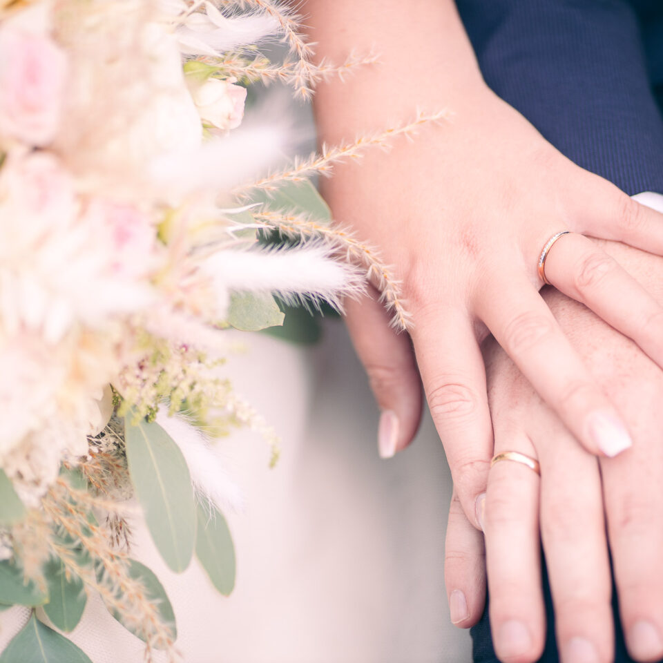 Detailaufnahme von zwei sich berührenden Händen mit Eheringen – im Vordergrund ein pastellfarbener Hochzeitsstrauß mit Rosen, Eukalyptus und Gräsern. Sanftes Licht und weiche Farben verleihen dem Bild eine zarte, emotionale Stimmung.