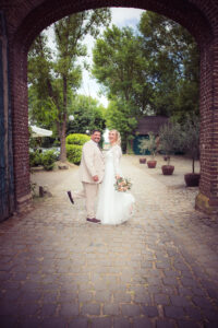 Lachendes Brautpaar unter dem Torbogen der Wasserburg Geretzhoven – die Braut im weißen Kleid mit Blumenstrauß, der Bräutigam hebt humorvoll das Bein, umgeben von Natur und rustikalem Charme.