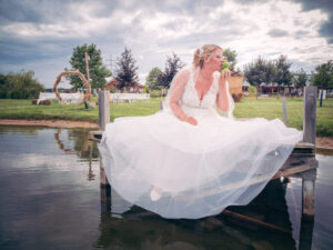 Braut in einem weißen Hochzeitskleid sitzt auf einem Holzsteg am Wasser und küsst mit einem Augenzwinkern eine grüne Froschfigur – freie Trauung im Hintergrund, märchenhafte Szene mit viel Humor.