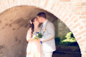 Hochzeitspaar küsst sich zärtlich unter einem alten Backsteingewölbe mit Brautstrauß in der Hand