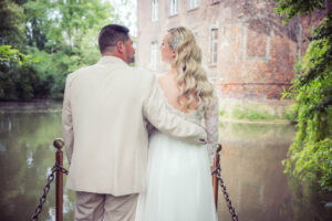 Braut und Bräutigam stehen Arm in Arm am Wasser vor einem alten Schloss und schauen sich liebevoll an