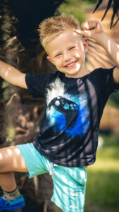Blondhaariger Junge mit Controller-T-Shirt lächelt und klettert am Baum – Sommerliches Kinderportrait
