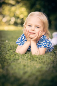 Ein blondes Mädchen im blauen Kleid mit Blümchenmuster liegt auf der Wiese, stützt den Kopf in die Hände und schaut verträumt in die Kamera. Weiches Licht und warmer Hintergrund sorgen für eine idyllische Stimmung.