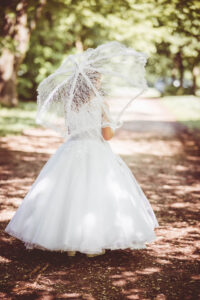 Kommunion Mädchen von hinten mit weißem Kleid und Spitze-Schirm auf einem Waldweg