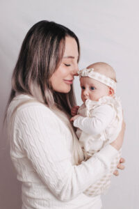 Eine Mutter hält ihr Baby liebevoll im Arm, beide tragen helle Kleidung vor neutralem Hintergrund