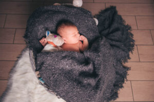 Ein Baby liegt nackt und eingekuschelt in eine dunkle Decke im Korb, festhaltend an einem Stofftier