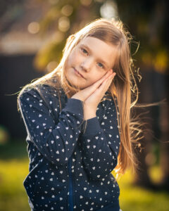Mädchen mit blonden Haaren legt den Kopf sanft auf die Hände – Kinderportrait im Garten bei warmem Gegenlicht