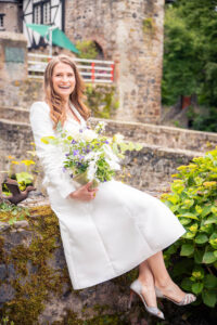 Die Braut sitzt auf einer Mauer vor historischem Gemäuer, mit einem frischen Sommerstrauß in der Hand