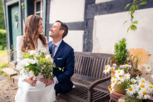 Brautpaar sitzt auf einer alten Holzbank vor Fachwerk, beide strahlen sich verliebt an