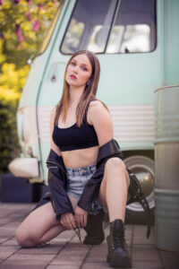 Ein junges Model posiert selbstbewusst im Streetstyle-Look mit Lederjacke, Crop-Top und Boots vor einem mintfarbenen Retro-Van. Die Pose ist ausdrucksstark, das Licht warm und kontrastreich.