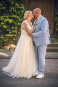 Ein Brautpaar küsst sich innig auf der Straße vor einer alten Holztür. Die tätowierte Braut trägt ein cremefarbenes Brautkleid, der Bräutigam ein hellblaues Sakko mit Sneakern. Authentischer Hochzeitsmoment voller Stil und Liebe.